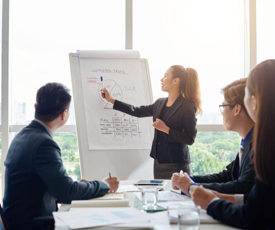 Attractive Asian businesswoman with ponytail pointing at diagram on marker board while holding working meeting in spacious boardroom