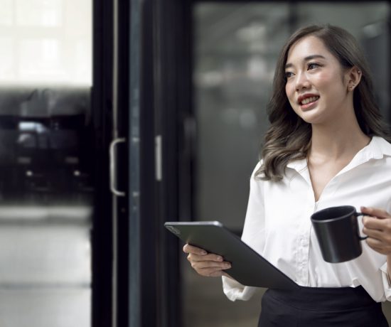 Portrait of young smiley asian businesswoman holding tablet and mug standing in modern officer.
